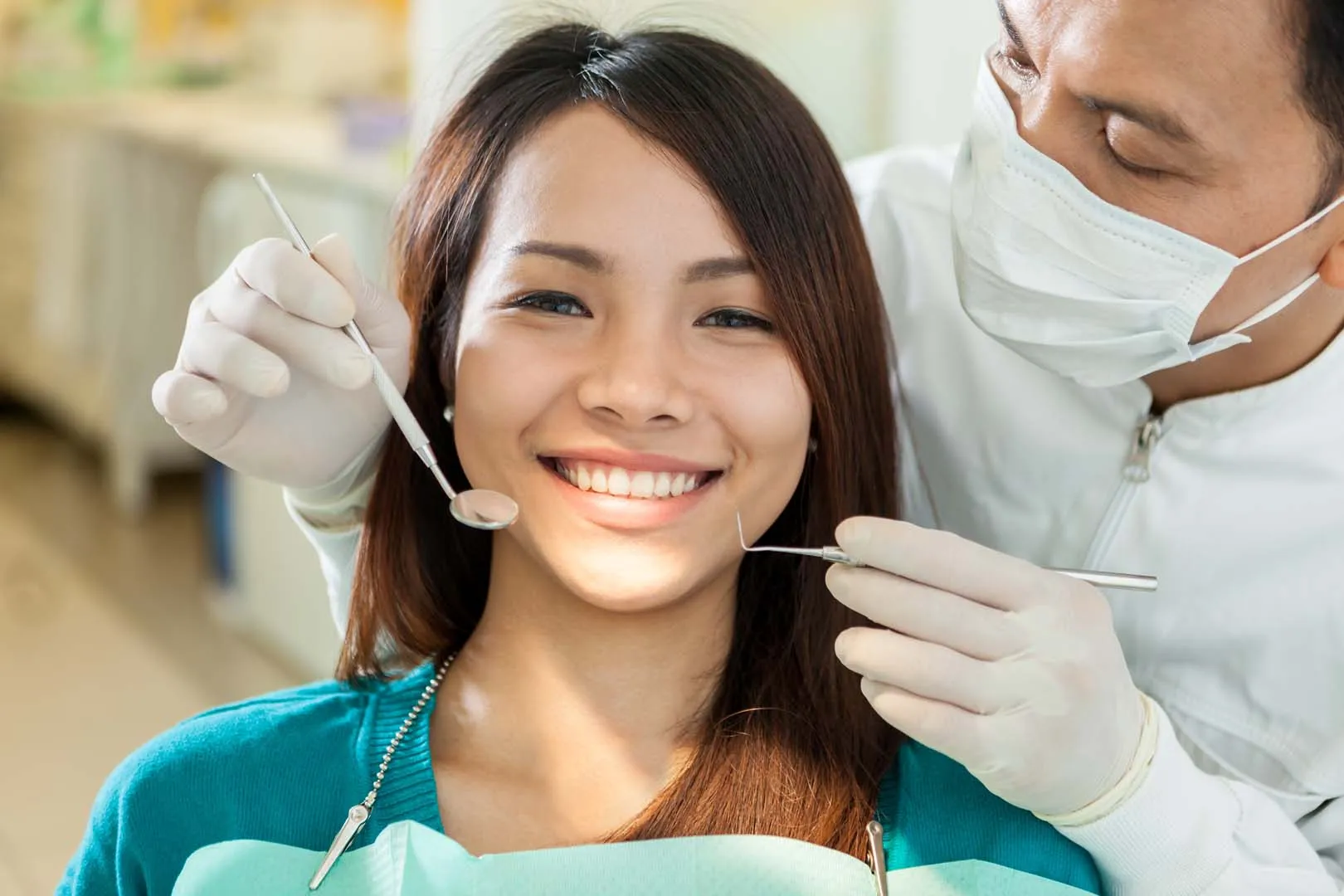 Dentist wearing mask and gloves examining smiling patient's teeth with dental tools.