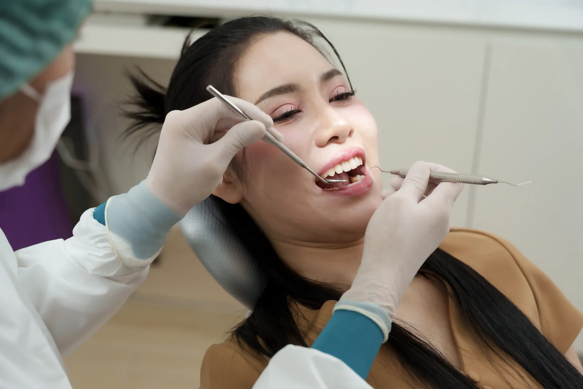 Dentist wearing gloves examines a woman's teeth with dental tools in a clinic chair.